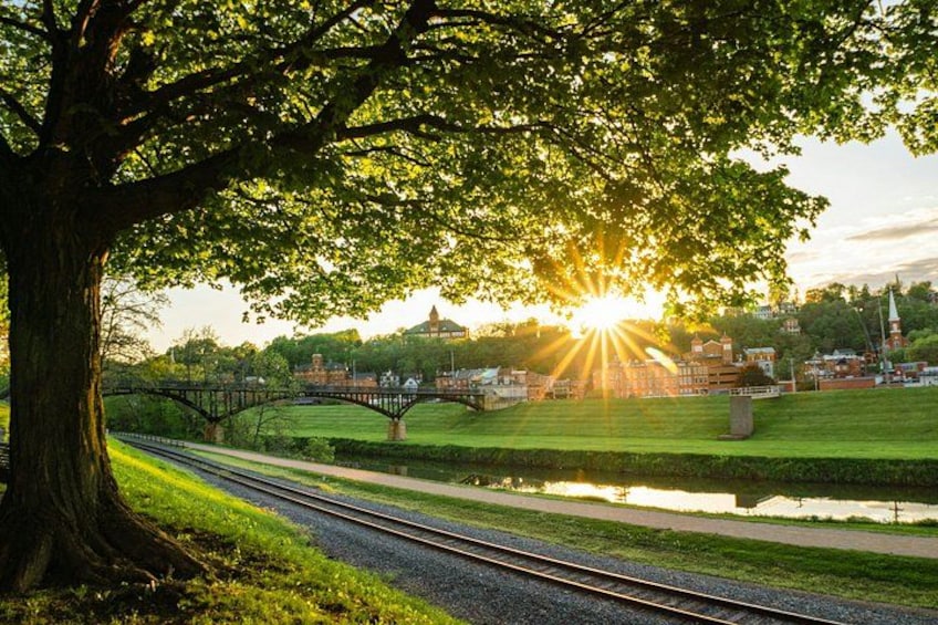 Een spoorlijn met bomen langs de kant, een brug en een zonnig landschap met gebouwen op de achtergrond. 5