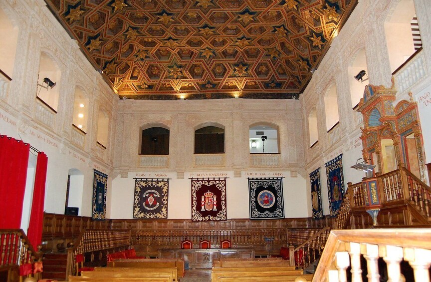 An ornate hall with a patterned ceiling, wooden pews, and banners with heraldic symbols. 5