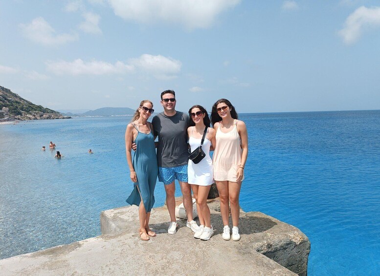 A group of people standing on a concrete ledge by a clear blue sea with distant hills and a few swimmers in the water. 1