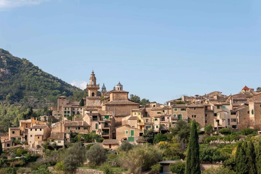 A historic village with terracotta-roofed buildings, a prominent church tower, and lush greenery. 1