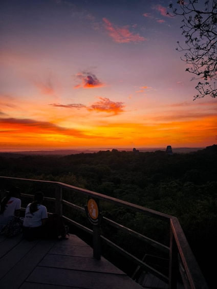 A sunset over a mountainous landscape with a wooden viewing platform and a sign indicating a hiking trail. 1