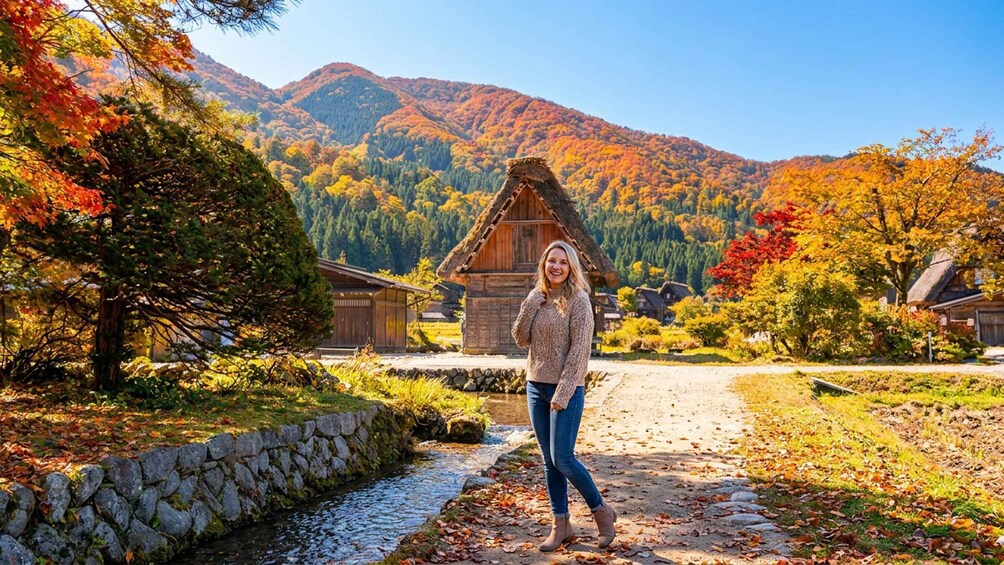 A woman standing on a stone path in a rural village with traditional wooden houses and autumn foliage. 5