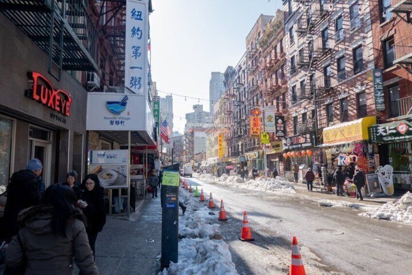 A snowy urban street with shops, a noodle restaurant, and orange traffic cones. 1