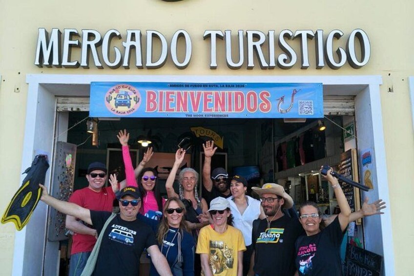 Un gruppo di persone all'ingresso di un mercato turistico, con un cartello che riporta 'MERCADO TURISTICO' e 'BIENVENIDOS'. 2