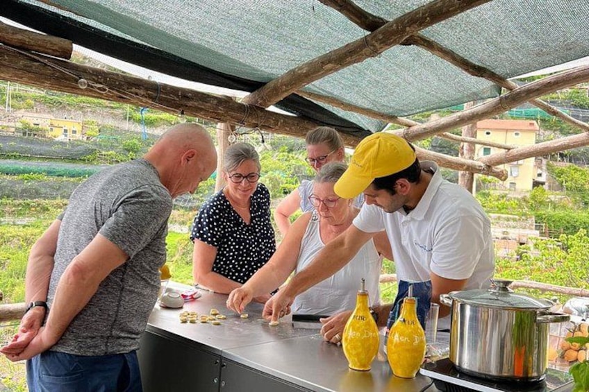 En gruppe mennesker samlet omkring et bord med kogegrej og flasker, under et overdækket område med udsigt til grønt og bygninger i baggrunden. 1