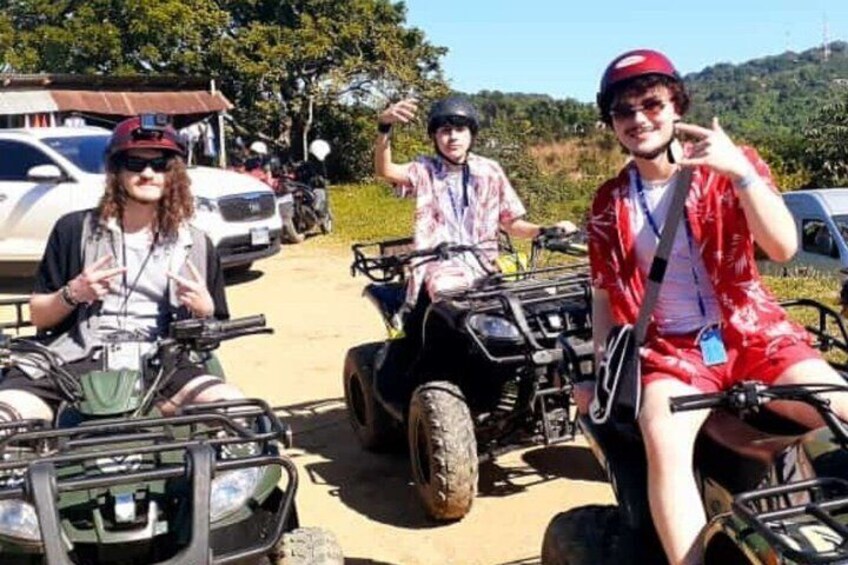 Three people on ATVs in a rural setting with vehicles and trees in the background. 4
