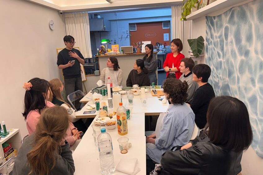 Un groupe de personnes est assis autour d’une table dans une pièce dont un mur est bleu et présente une texture particulière, et on aperçoit un coin cuisine à l’arrière-plan. 5