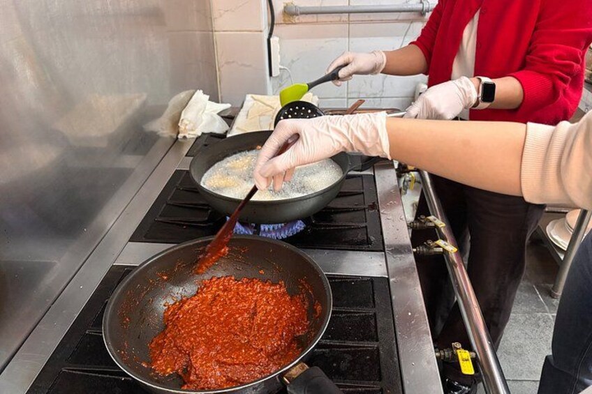 Une personne cuisine dans une poêle sur une cuisinière, une autre personne remue le contenu d’une casserole, et une grande terrine contient de la sauce. 4