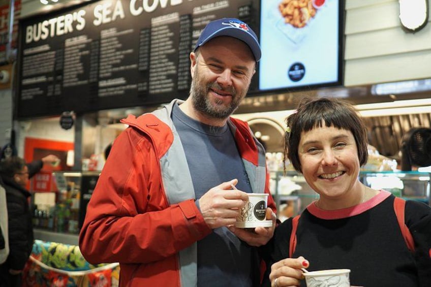 A person in a red jacket holding a cup, standing in front of a menu board with the text 'BUSTER'S SEA COVE'. 1