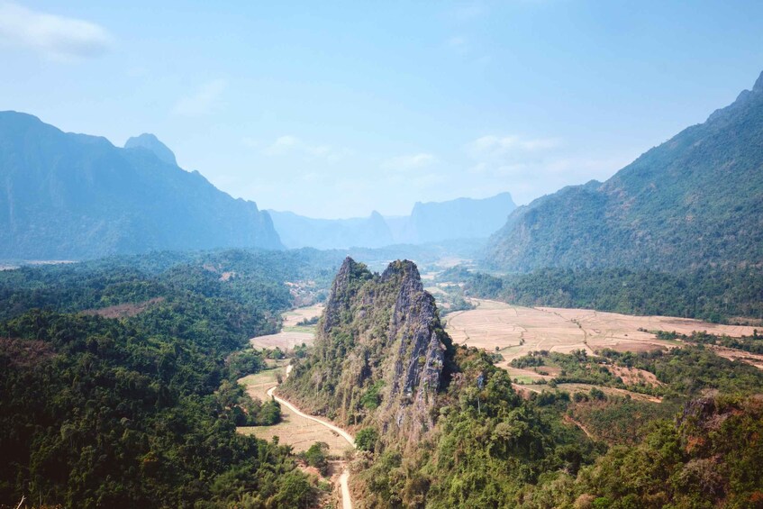 A mountainous landscape with a prominent rocky peak, a winding road, and surrounding forested areas. 3
