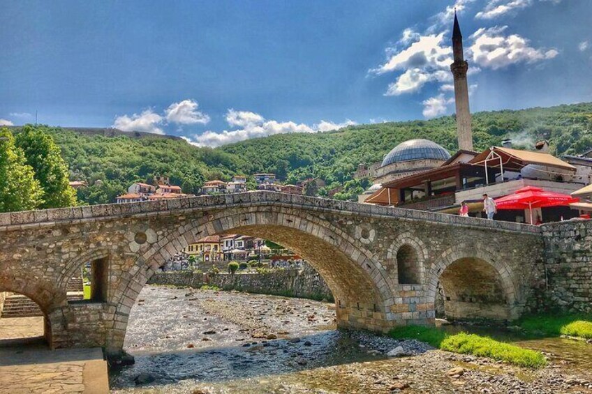 A stone bridge over a river, a mosque with a minaret, and a dome in a hilly town. 1