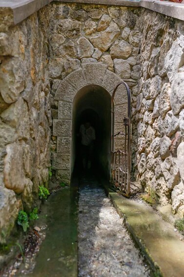 A stone tunnel with a metal gate, water flowing on the ground, and a person walking inside. 4
