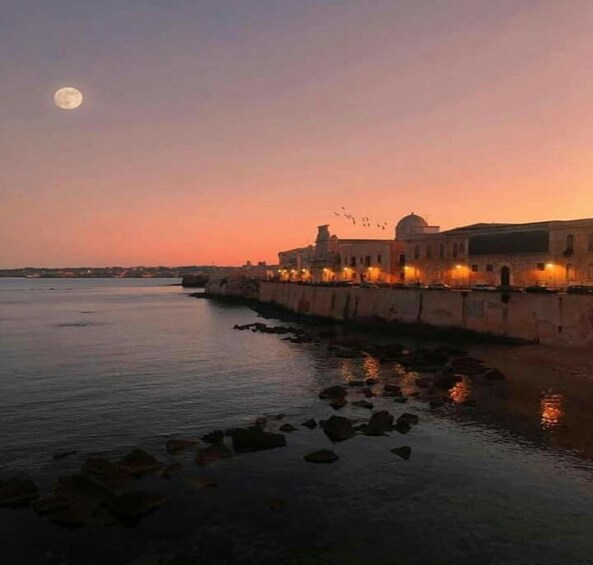 A coastal cityscape at dusk with a prominent building featuring a dome, illuminated windows, and a calm sea with rocks in the foreground. 3