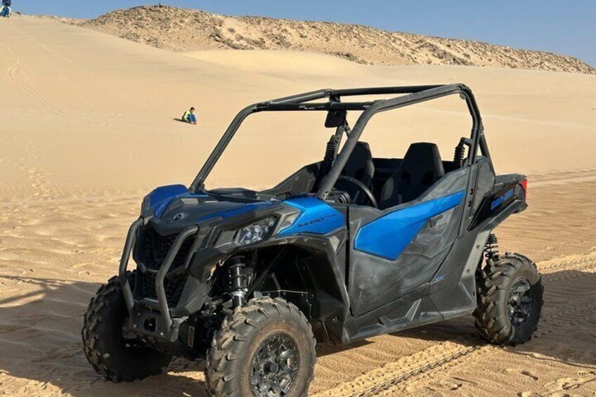 Un veicolo fuoristrada blu e grigio parcheggiato su un terreno desertico sabbioso, con una persona visibile in lontananza. 2