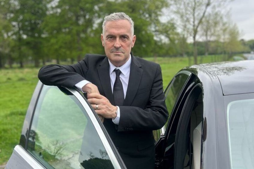 A man in a suit leaning out of a car window in a grassy area. 3