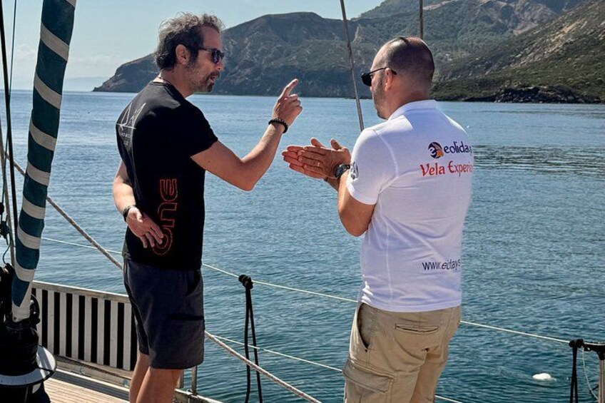 Two people on a boat, one wearing a black t-shirt and the other in a white t-shirt with a logo, standing by a calm body of water with hills in the background. 3
