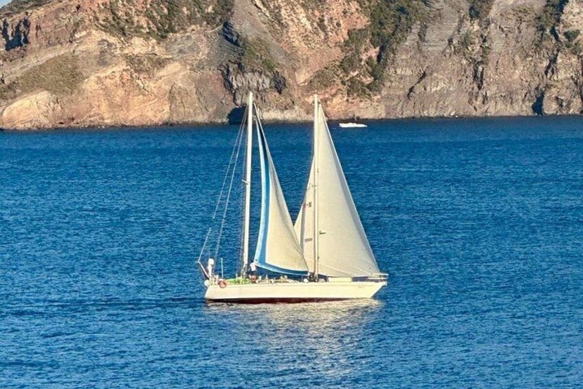 A sailboat with a white hull and blue sails on a calm sea with rocky cliffs in the background. 1