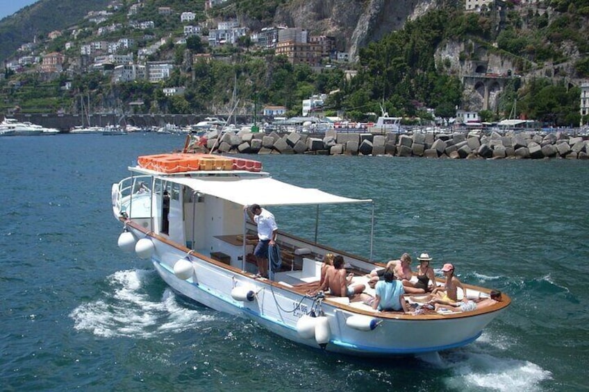 A boat with passengers on a calm sea, a rocky coastline, and buildings in the background. 2