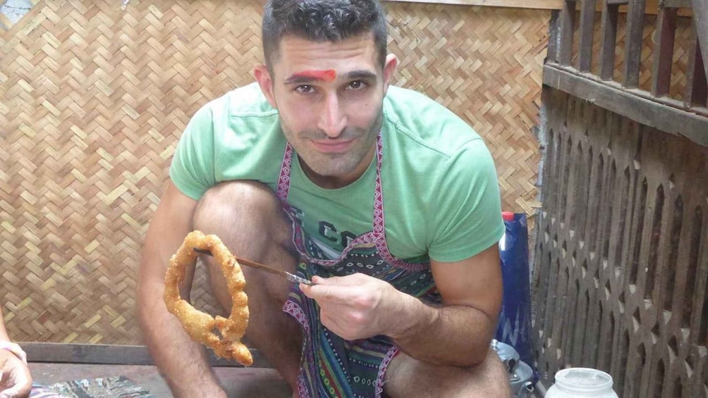 A person holding a fried food item outdoors, with a woven fence and a metal container in the background. 4