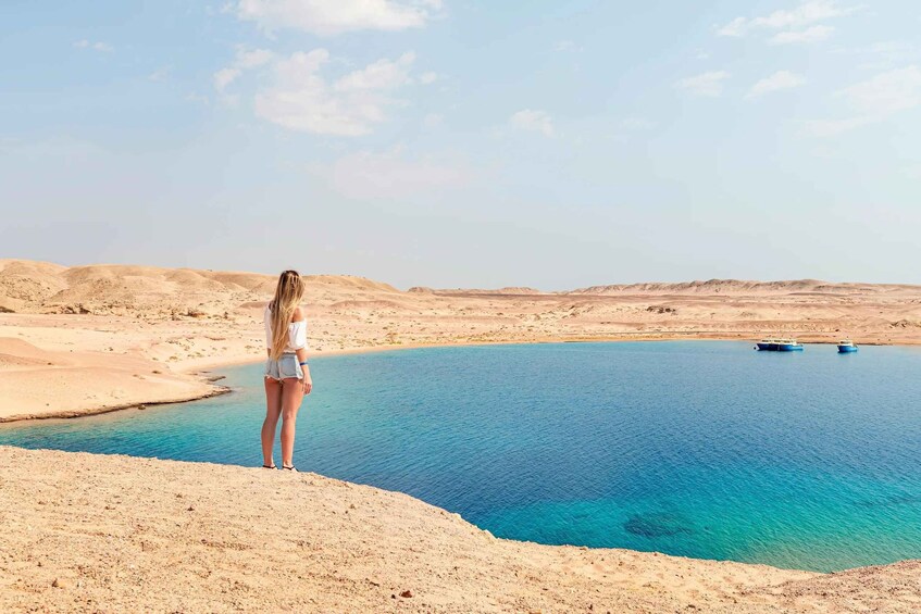 A person standing on a sandy beach overlooking a calm blue lake with boats in the distance. 1