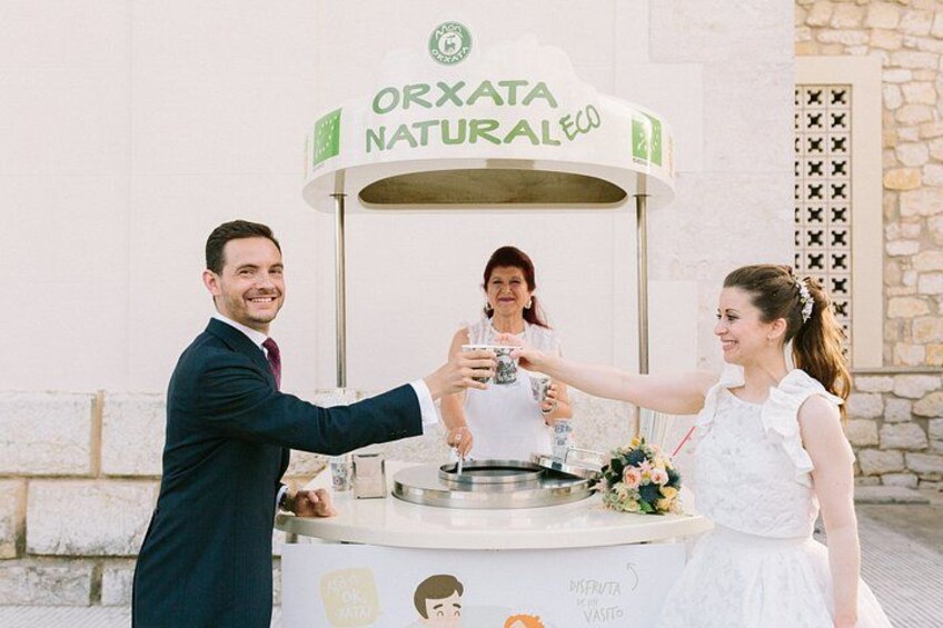 Eine Hochzeitszeremonie, auf der sich ein Paar erfreut anstoßt und ein Kuchen mit der Aufschrift 'ORXATA NATURALECO' vor einem Steinhaus steht. 5