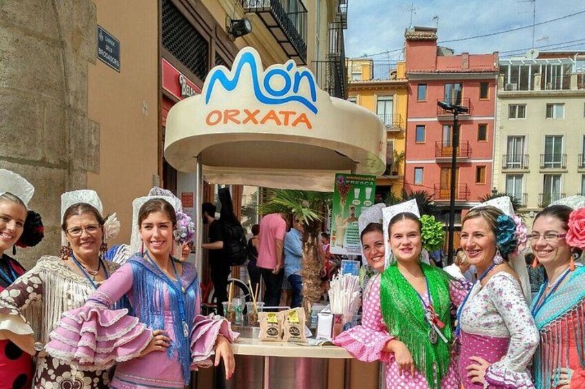 Eine Gruppe von Menschen in traditioneller Kleidung steht vor einem Essensstand mit einem Schild, auf dem 'Mon ORXATA' steht. 2