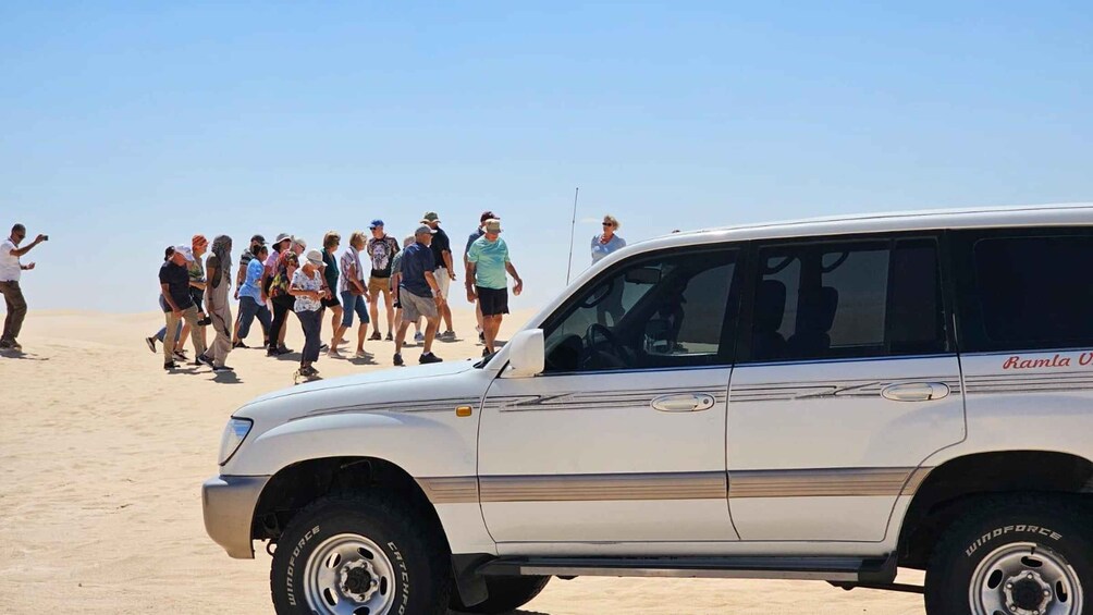 A white SUV with 'Ramla U' on the side parked on a sandy beach with a group of people in the background. 2