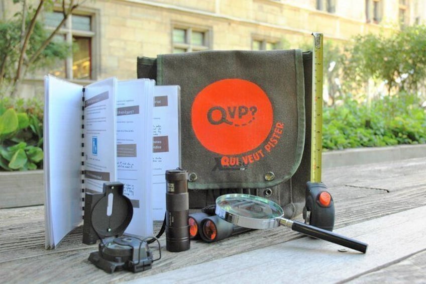 A collection of items including a notebook, a magnifying glass, a measuring tape, and a bag with 'OVP?' on it, placed on a wooden surface outdoors. 5