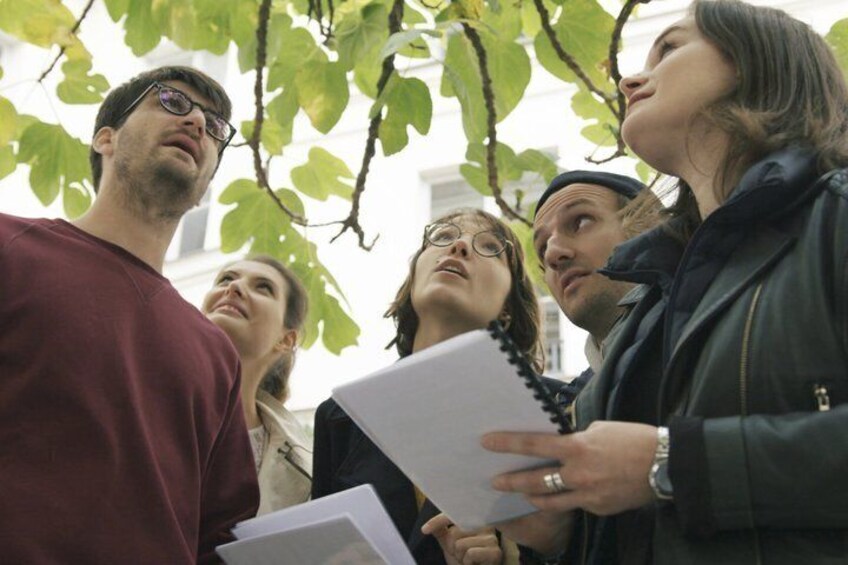A group of people standing outdoors, some holding papers, under a tree with green leaves. 2