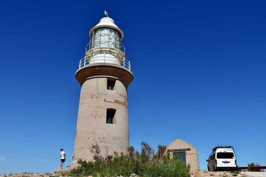 A lighthouse with a person standing nearby and a vehicle parked in front. 4