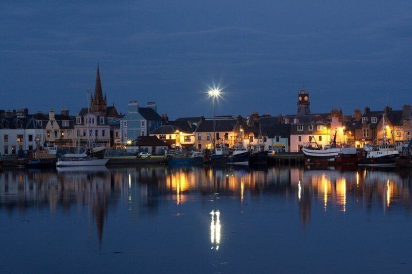 A harbor at dusk with buildings reflecting on the water, a prominent church spire, and a lighthouse. 2
