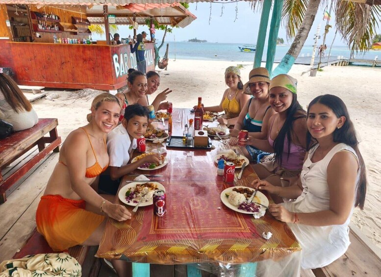 A group of people dining at a wooden table on a beach with a bar in the background. 4