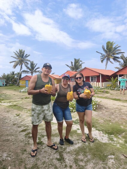 Three people holding pineapples in a tropical setting with palm trees and red-roofed buildings. 2