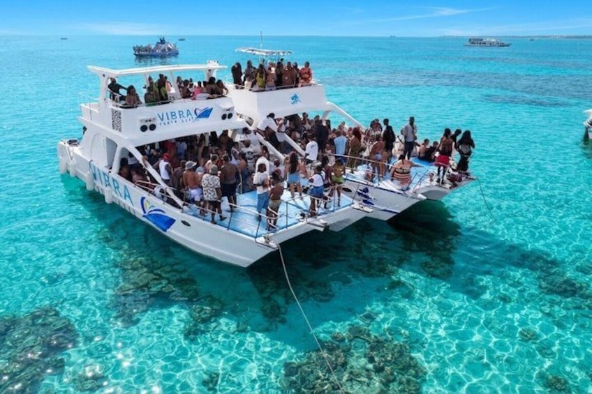A boat named 'VIBRA' with passengers on deck, surrounded by clear turquoise water and visible underwater formations. 3