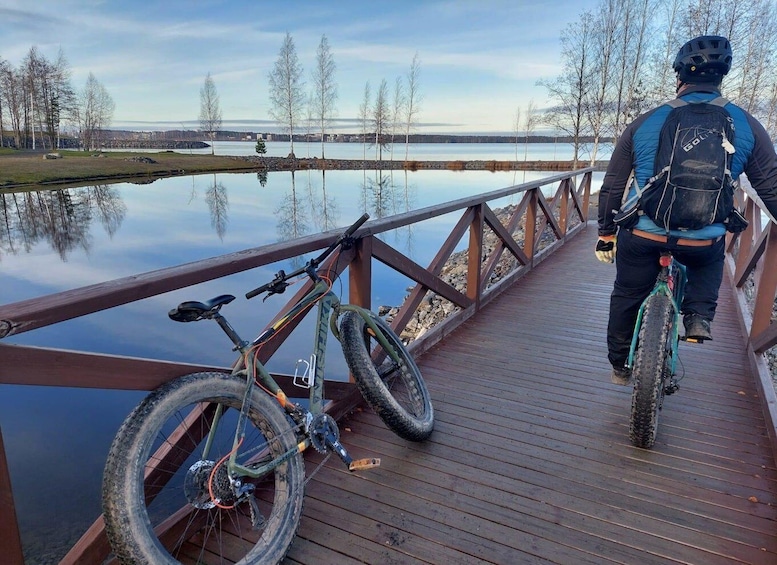 A person on a wooden bridge with a bicycle and a backpack, overlooking a calm lake with trees. 2
