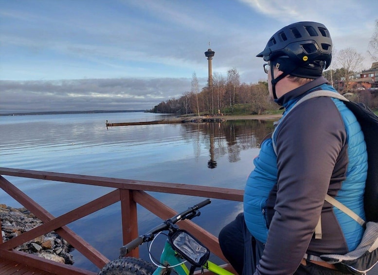 A person sitting on a dock with a bicycle, overlooking a calm lake with a tower in the distance. 1
