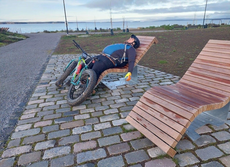 A person resting on a wooden bench with a mountain bike and gear beside them. 5