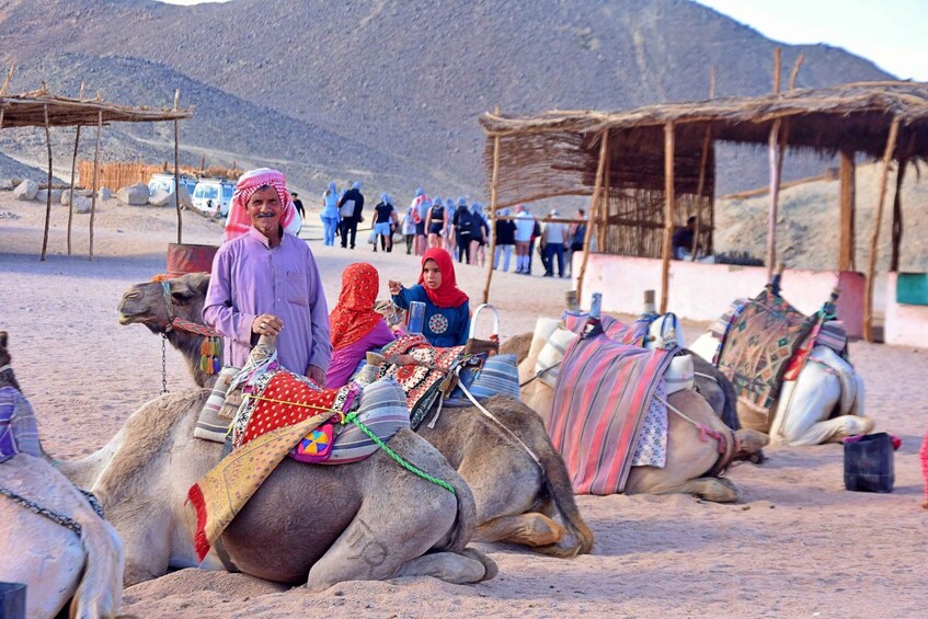 Camels with riders in a desert setting with tents and a mountainous backdrop. 1