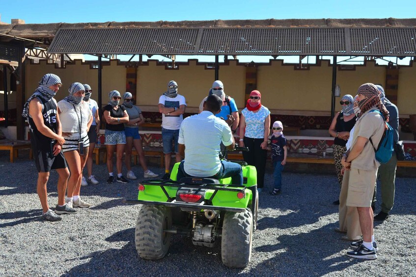 A group of people gathered around a green ATV in a rural setting with a thatched-roof building in the background. 4