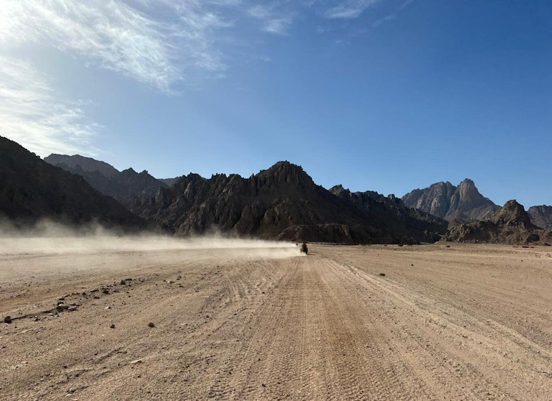 A desert landscape with a dirt road, a vehicle creating dust, and rugged mountains in the background. 2