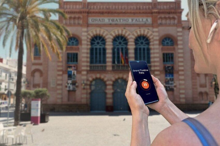 A person using a smartphone with a theater app, standing in front of the Grand Teatro Fallia. 5