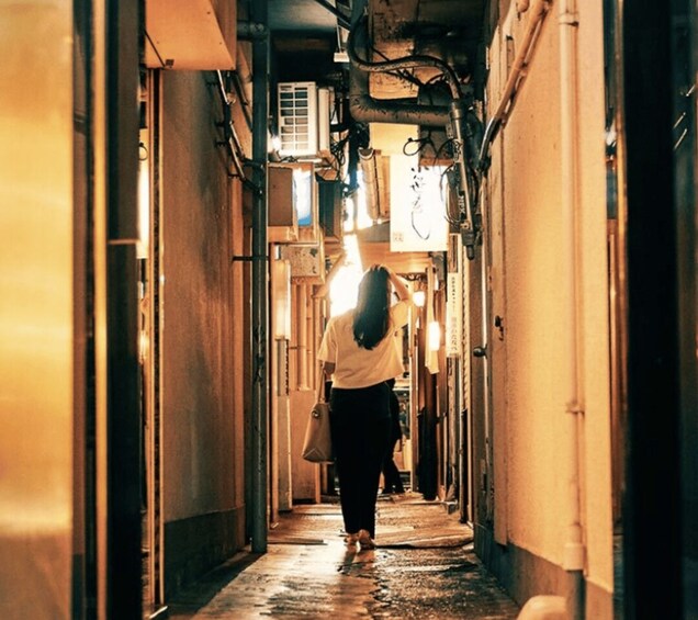 A person walking down a narrow corridor with overhead pipes and a window on the right. 3