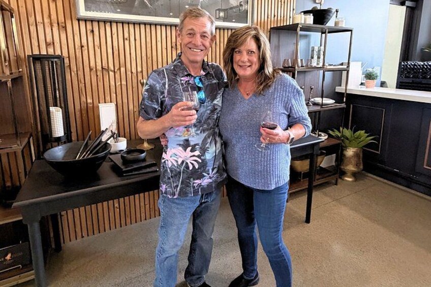 Two people standing in a kitchen with wooden paneling, holding glasses of wine. 4