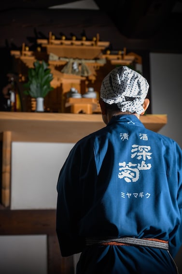 A person in a blue traditional garment with Japanese characters on the back, standing in a room with wooden furniture and a plant. 4