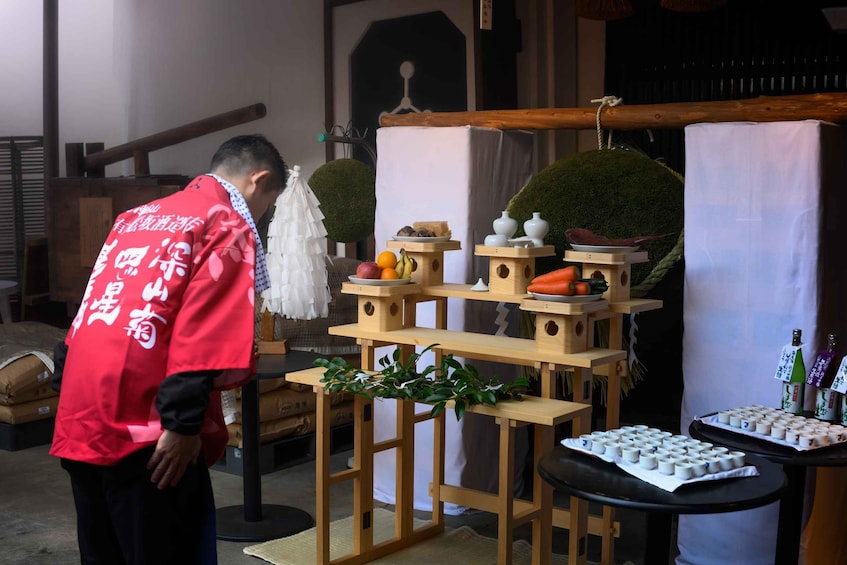 A person in a red shirt with white text standing in front of a wooden display with various items including fruits, vegetables, and bottles. 3