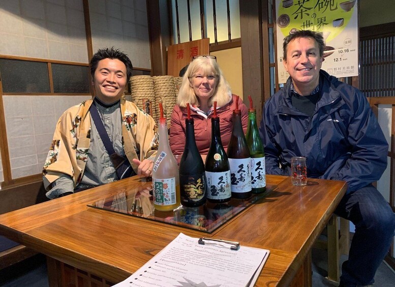 Three people seated at a wooden table with bottles of Japanese sake. 1
