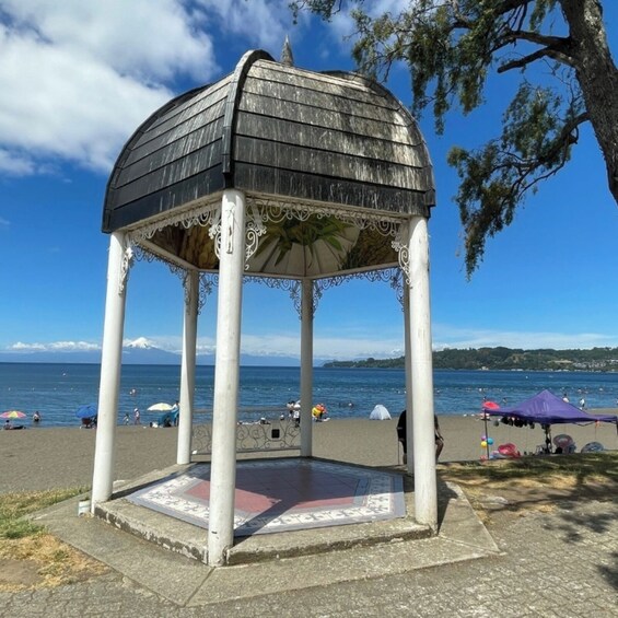En pavillon med et dekorativt tag og et mønstret gulv, beliggende på en strand med hav og bakker i baggrunden. 3