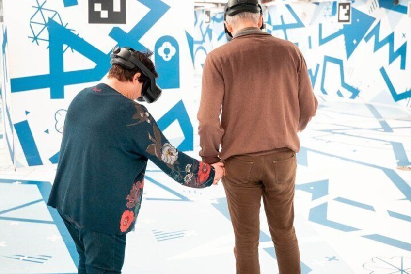Two people in a VR setup, one wearing a VR headset, in front of a wall with blue graffiti. 1