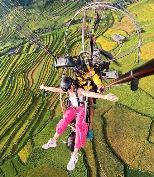 A person in a yellow and black harness with pink pants and white shoes, suspended in a cable car above terraced rice fields. 4