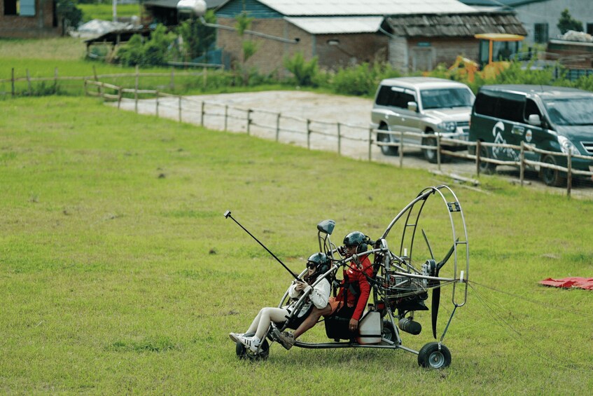 A person in a hang glider with a camera, a grassy field, and parked vehicles in the background. 3